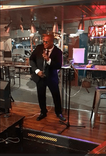 A bald man in a dark suit and sunglasses sings into a microphone on stage.